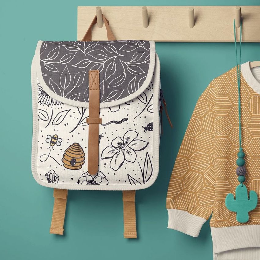 Kids backpack mockup hanging on a wooden peg rail, featuring a nature-themed line art design with bees, flowers, and leaves.