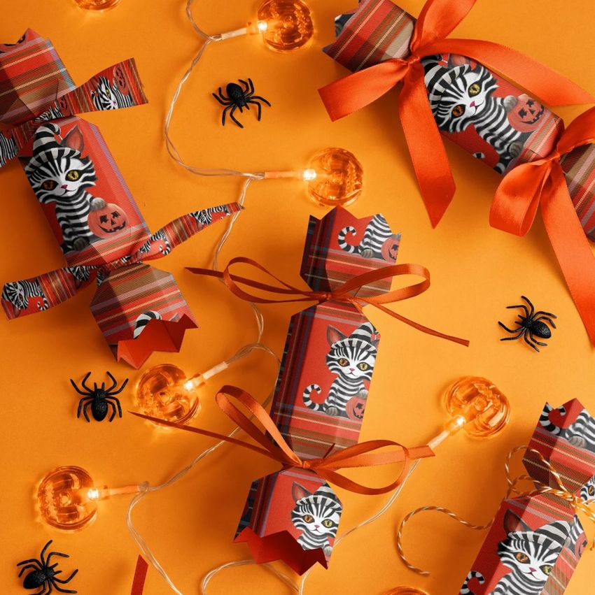 Halloween party cracker packaging mockup featuring striped cat and pumpkin designs on orange and red patterned paper, surrounded by string lights and plastic spiders.