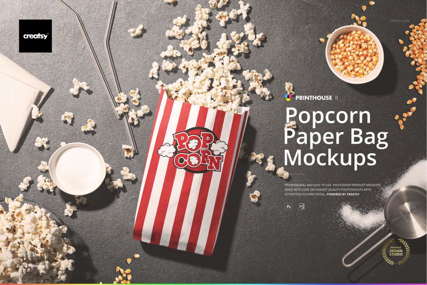 Top view of striped popcorn paper bag mockup with loose popcorn, bowl, and spoon on a dark textured surface.