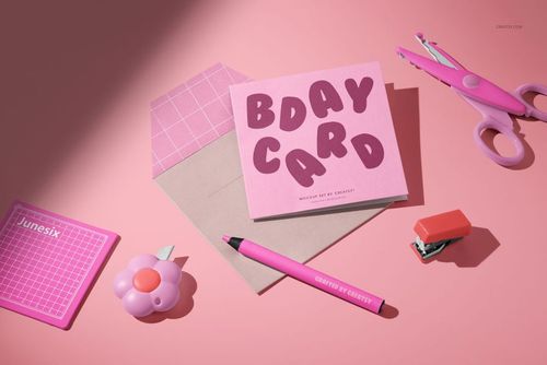 PSD mockup of a square greeting card on a pastel pink background with envelope, pen, scissors, and stapler props.