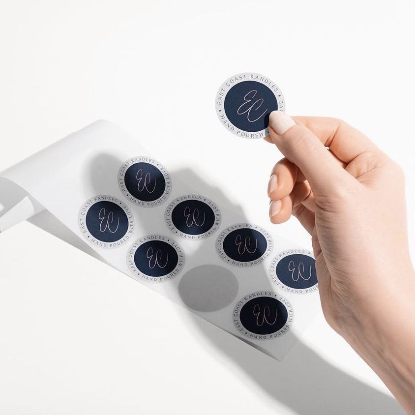 Hand holding a round sticker above a sheet of similar stickers on a white surface, with dark backgrounds and light script text.