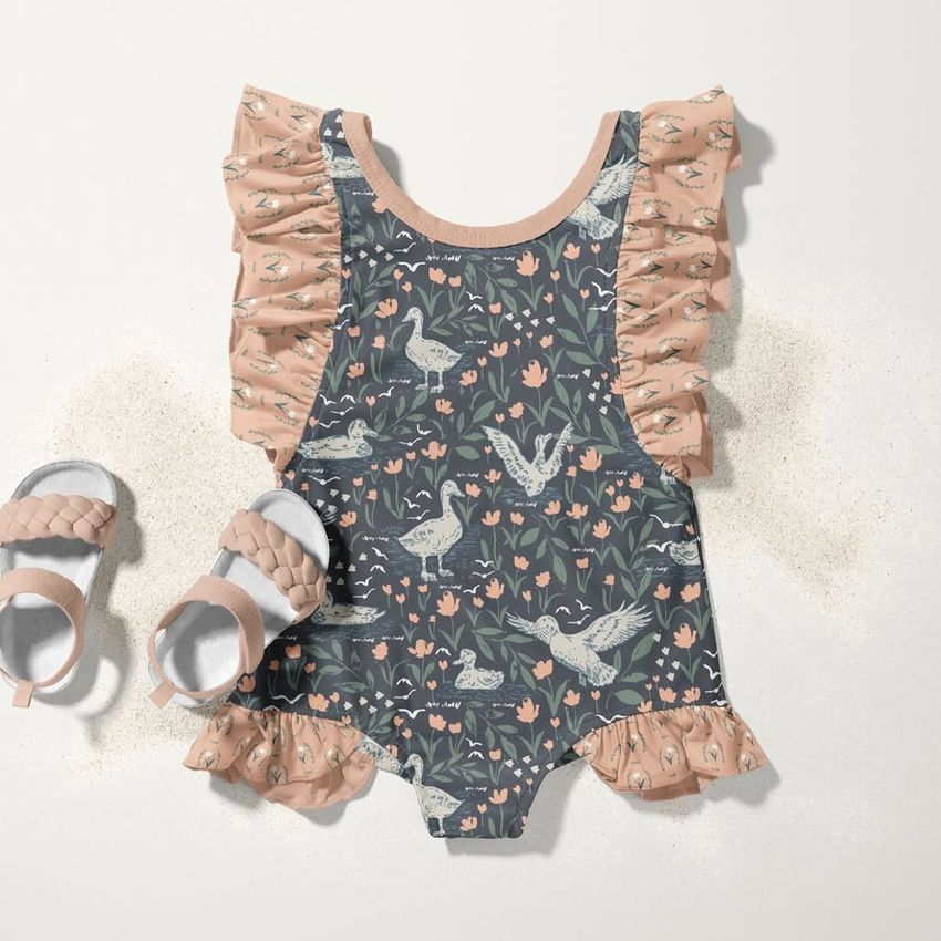 Baby ruffle swimsuit mockup featuring a dark floral and bird pattern with pink ruffled edges, displayed beside woven baby sandals on a light surface.