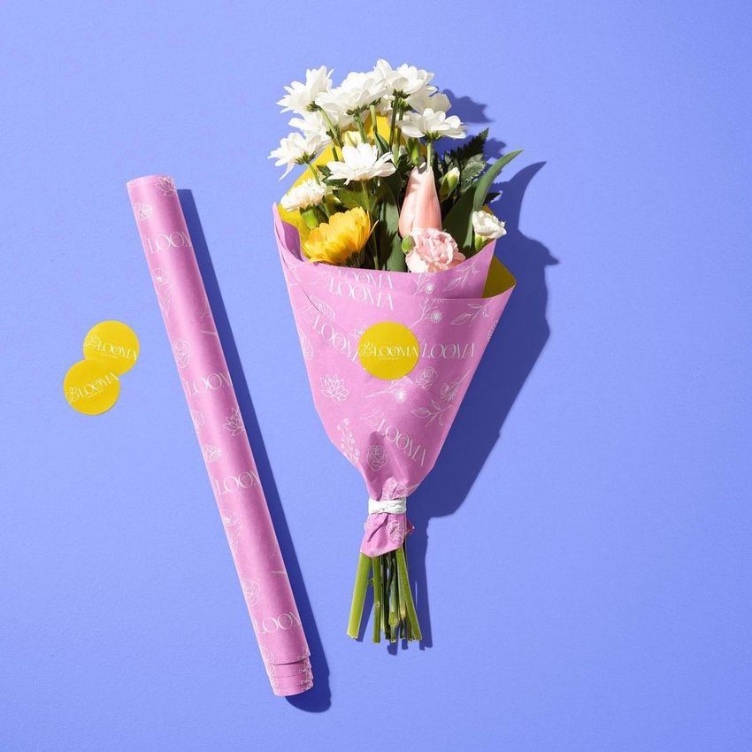 Flowers packaging mockup featuring a bouquet wrapped in pink floral paper, a matching roll, and two yellow stickers on a blue background.