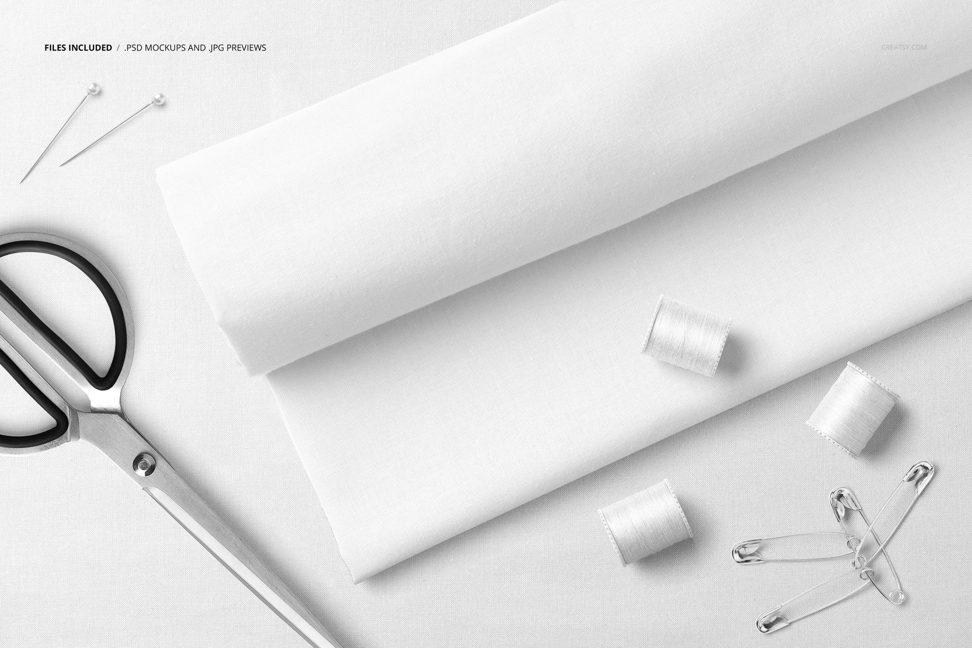 Flat lay of white cotton fabric mockup with sewing accessories including scissors, thread spools, pins, and safety pins.