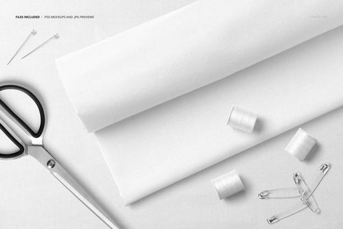 Flat lay of white cotton fabric mockup with sewing accessories including scissors, thread spools, pins, and safety pins.