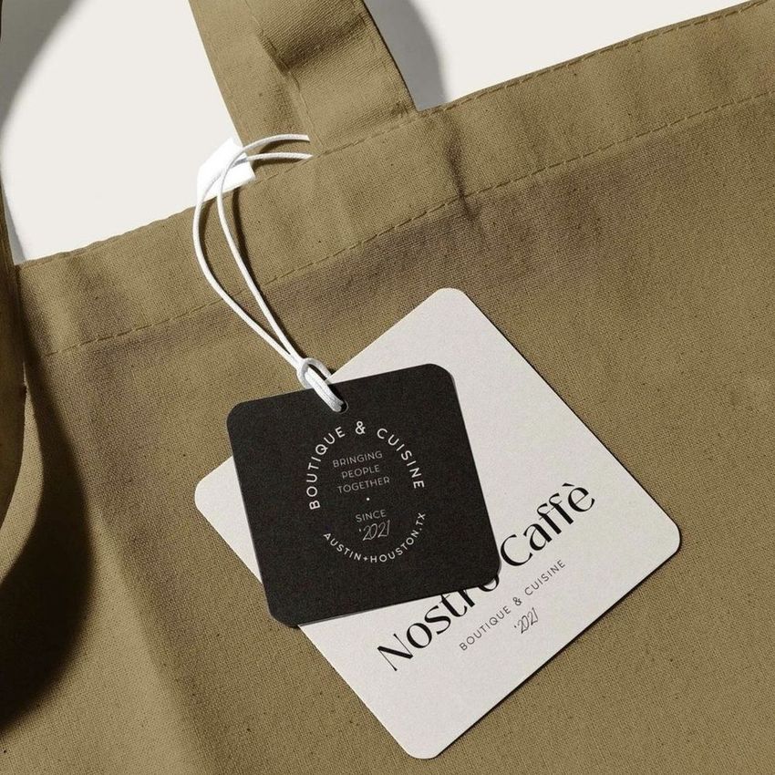 Canvas tote bag mockup showing a close-up of the handle and two attached tags, one black and one white, on a neutral background.