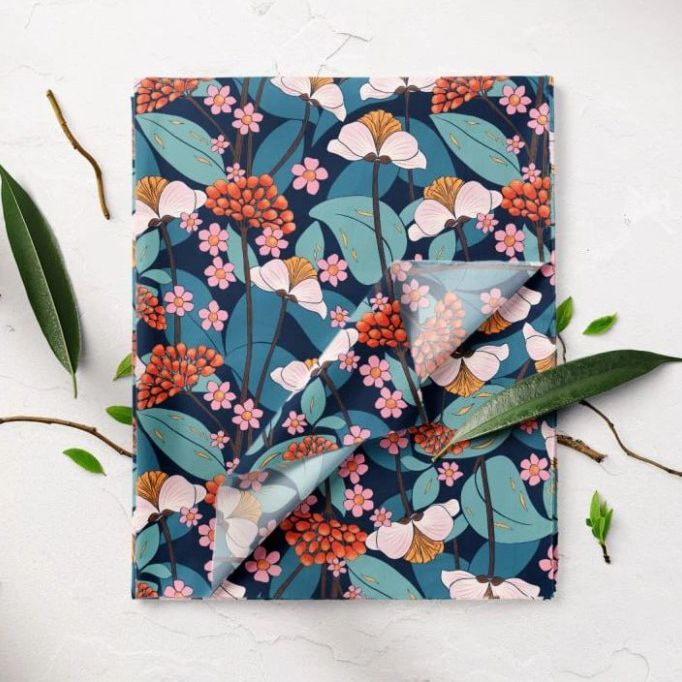 Folded fabric mockup featuring a floral pattern with pink, orange, and white flowers on a dark blue background, shown on a light surface with green leaves and twigs.