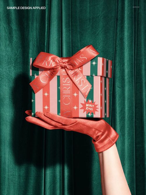 Christmas PSD mockup showing a hand in a shiny red glove holding a decorated gift box with bow, set against green drapes.