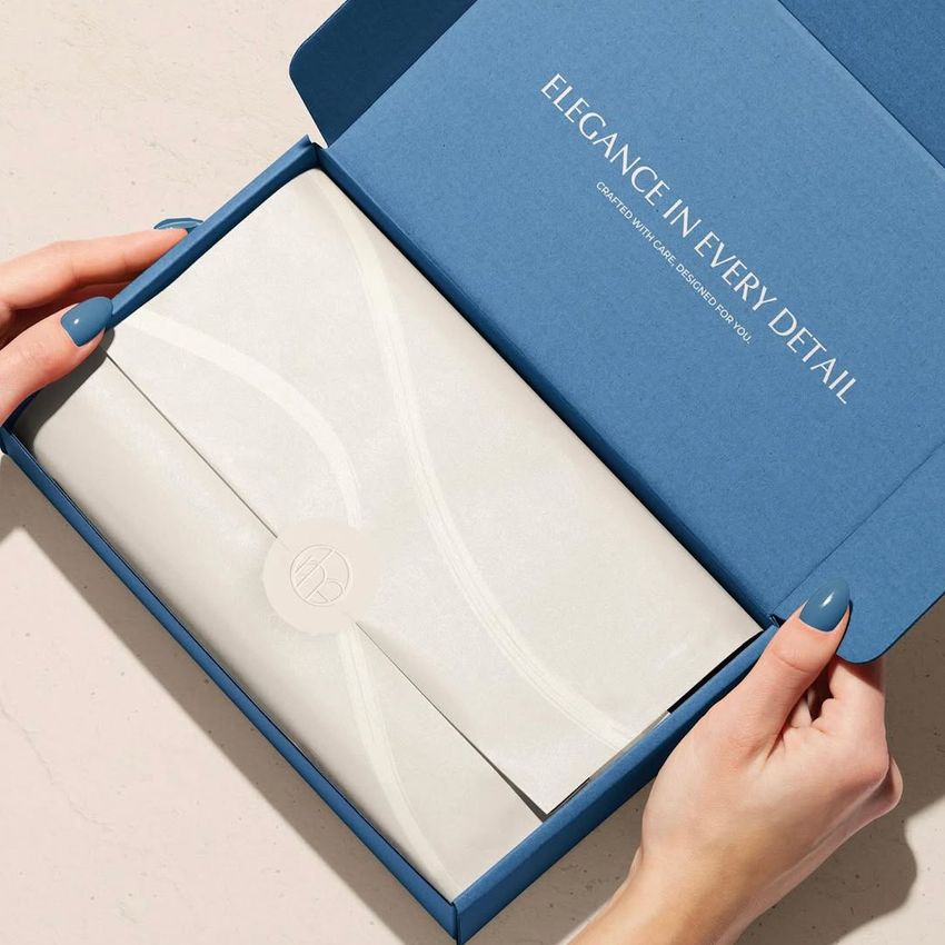 A person opens a blue mailing box mockup, revealing white tissue-wrapped contents, on a neutral tabletop background.