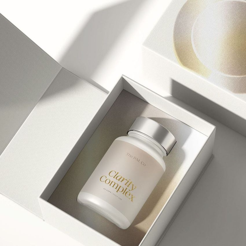 Cosmetics box packaging mockup featuring a white capsule bottle with gold text, placed inside an open textured box on a light surface.