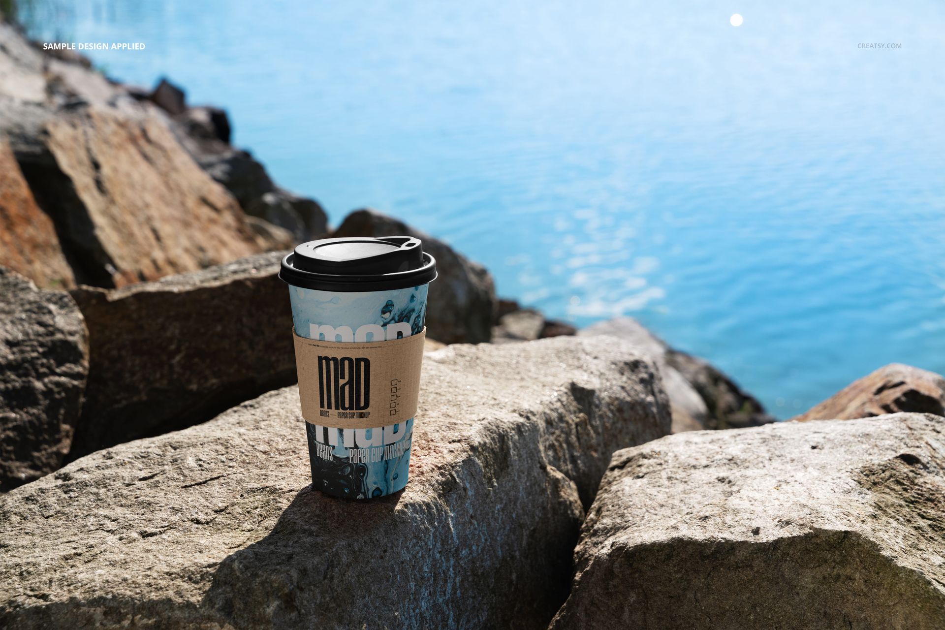 PSD mockup of a paper coffee cup with a black lid, blue pattern, and sleeve set on rocky shore near clear water.