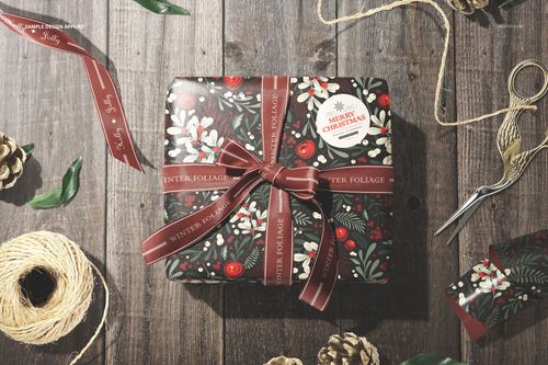 Wrapping paper mockup displaying a festive botanical print with ribbon, placed on rustic wood with crafting props.