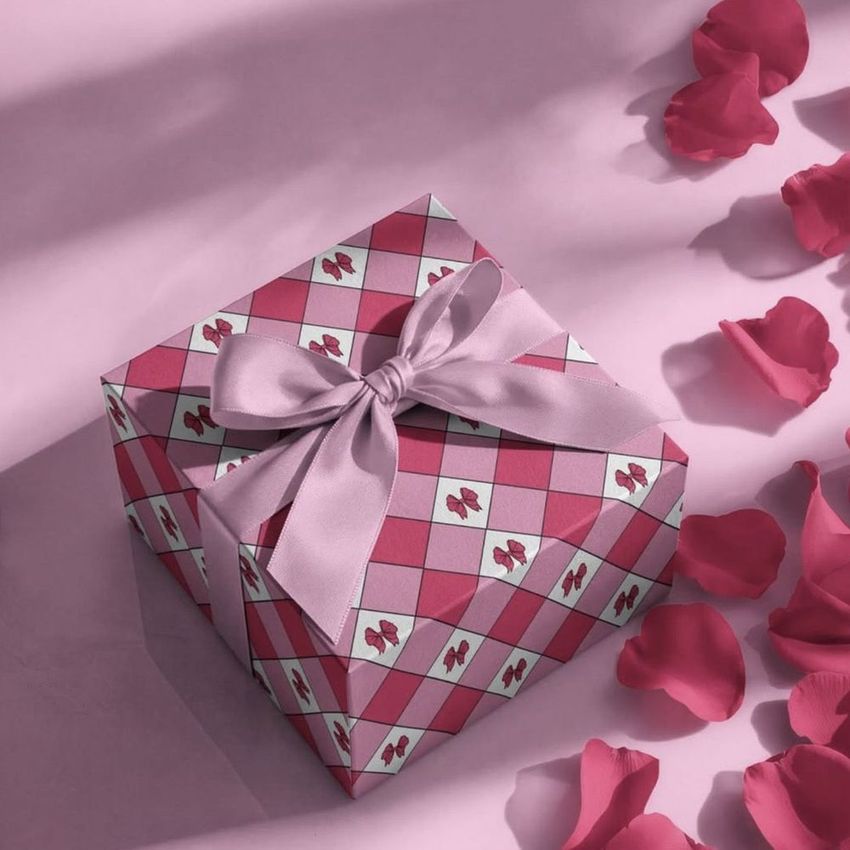 Square gift box mockup with a pink ribbon, decorated in a diamond pattern and surrounded by scattered rose petals on a pink surface.