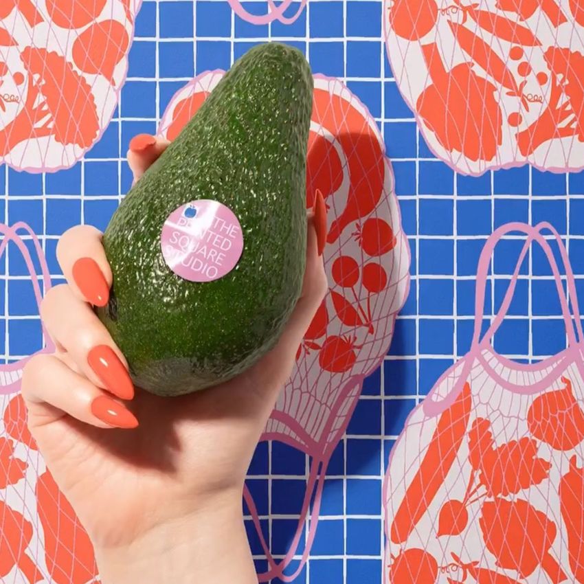Hand with orange nails holding an avocado with a round sticker, in front of a patterned fruit and vegetable wrapping mockup.