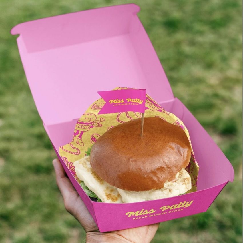 Hand holding a pink vegan burger packaging mockup with branded paper liner and flag, set against a grassy outdoor background.