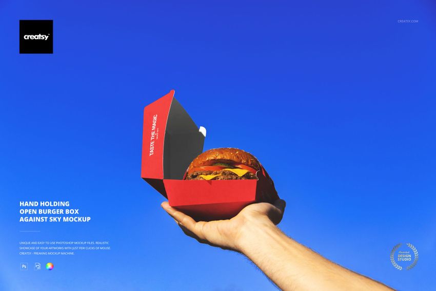 Hand holding an open red burger box with a hamburger inside against a clear blue sky, front view mockup.