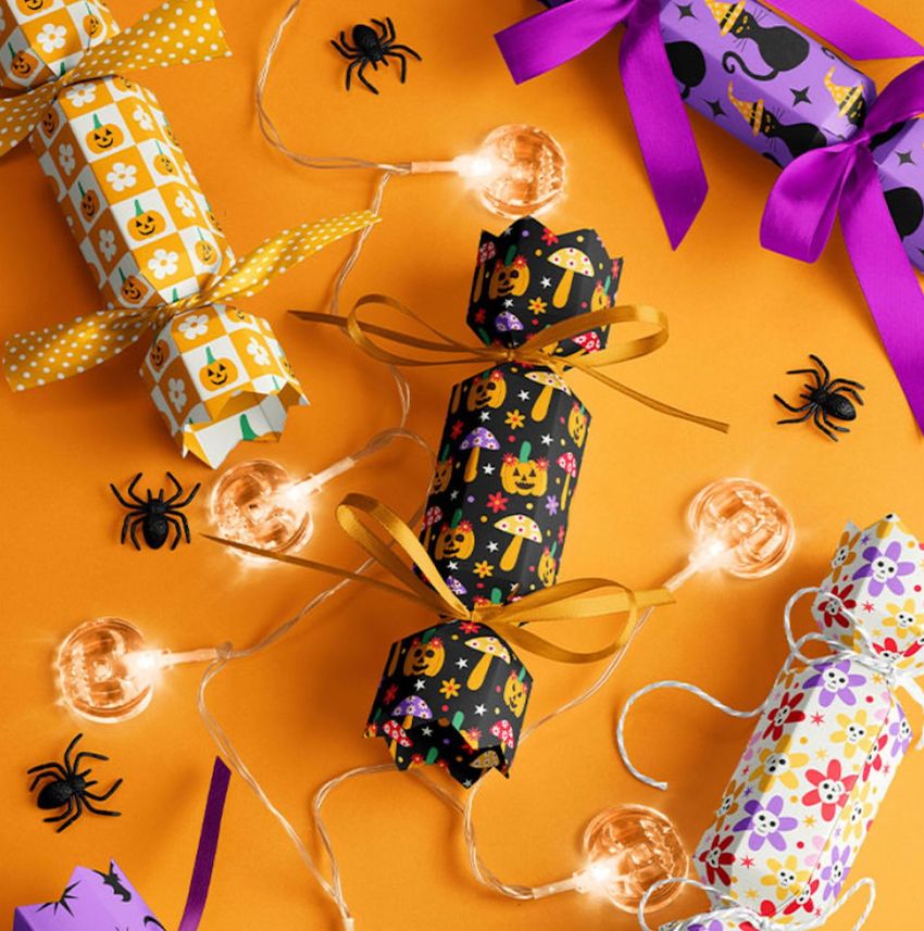 Halloween party cracker packaging mockup with colorful patterned crackers, string lights, and plastic spiders on an orange background.