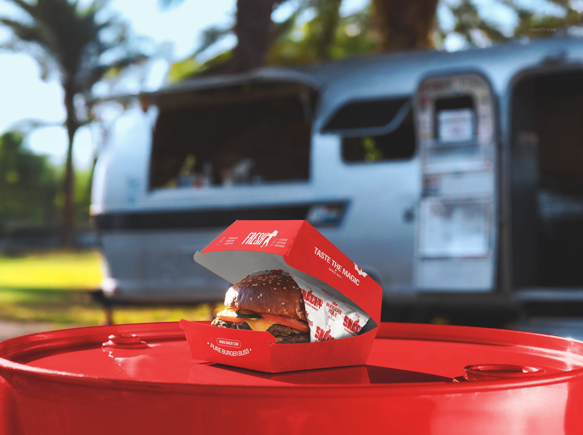 Fast food PSD mockup showing a burger in a red box on a barrel, outdoor scene with trailer and greenery behind.
