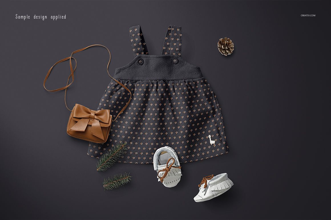 Front view of a baby dress mockup with a dark patterned skirt, brown purse, baby shoes, pine sprig, and pine cone on a dark background.