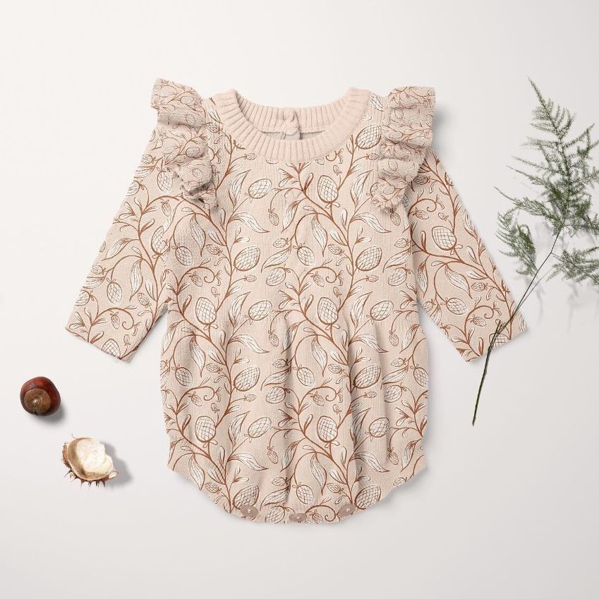 Knitted romper mockup featuring a beige baby romper with a brown botanical pattern, ruffled shoulders, and long sleeves on a light background with a sprig of greenery and two chestnuts.