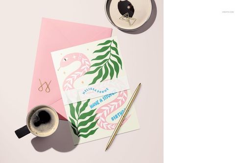 A5 card mockup with a pink envelope, gold pen, coffee cup, and ceramic dish on a light desk surface.