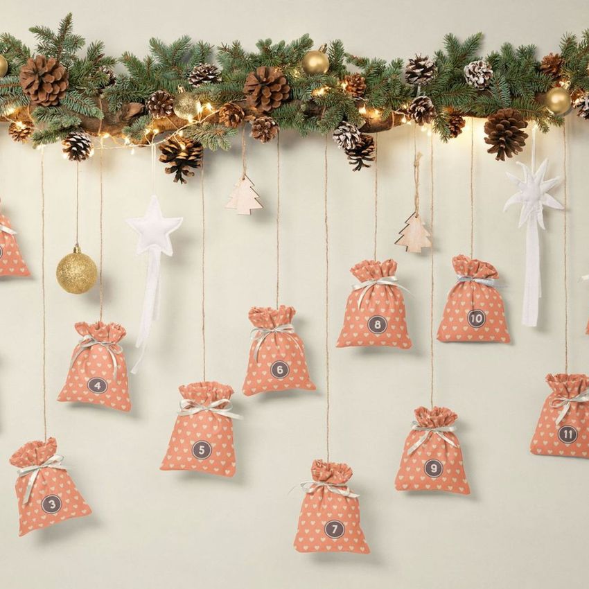 Drawstring sacks advent calendar mockup featuring small beige bags with heart patterns, numbered and hanging from twine under a pine garland with pinecones and ornaments.