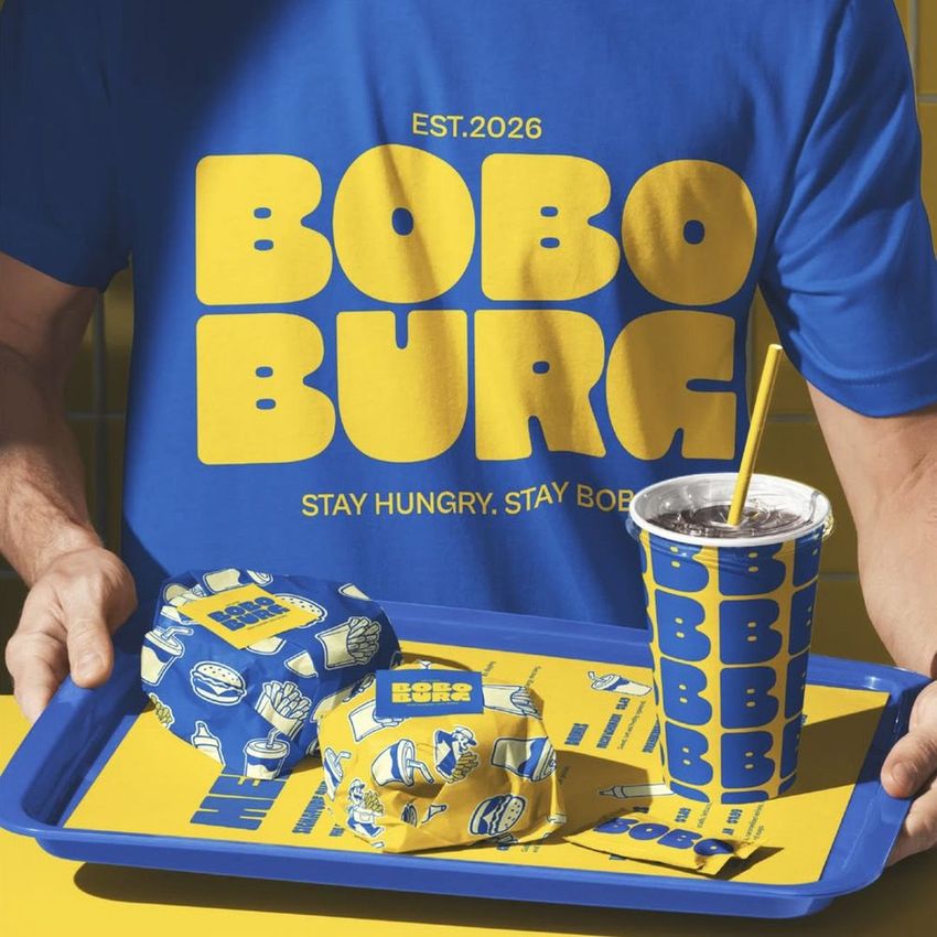 Man holding a tray with wrapped burgers and a drink cup, all featuring bold yellow and blue branding in a restaurant mockup.