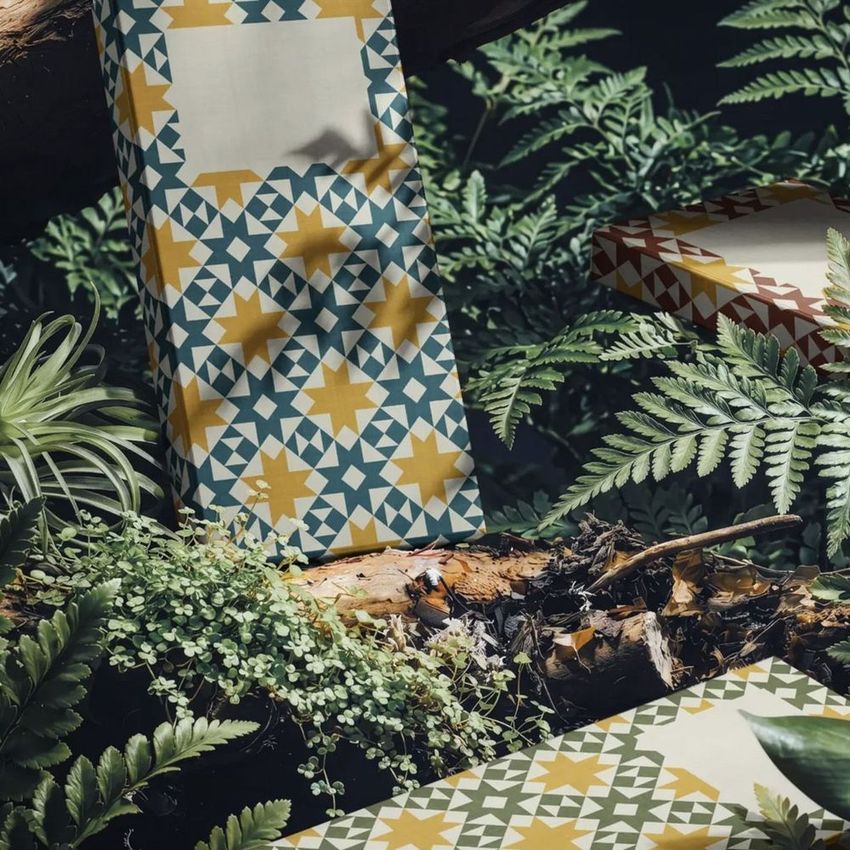 Chocolate box mockup with geometric quilt-inspired patterns in teal, yellow, and brown, placed among lush forest greenery.