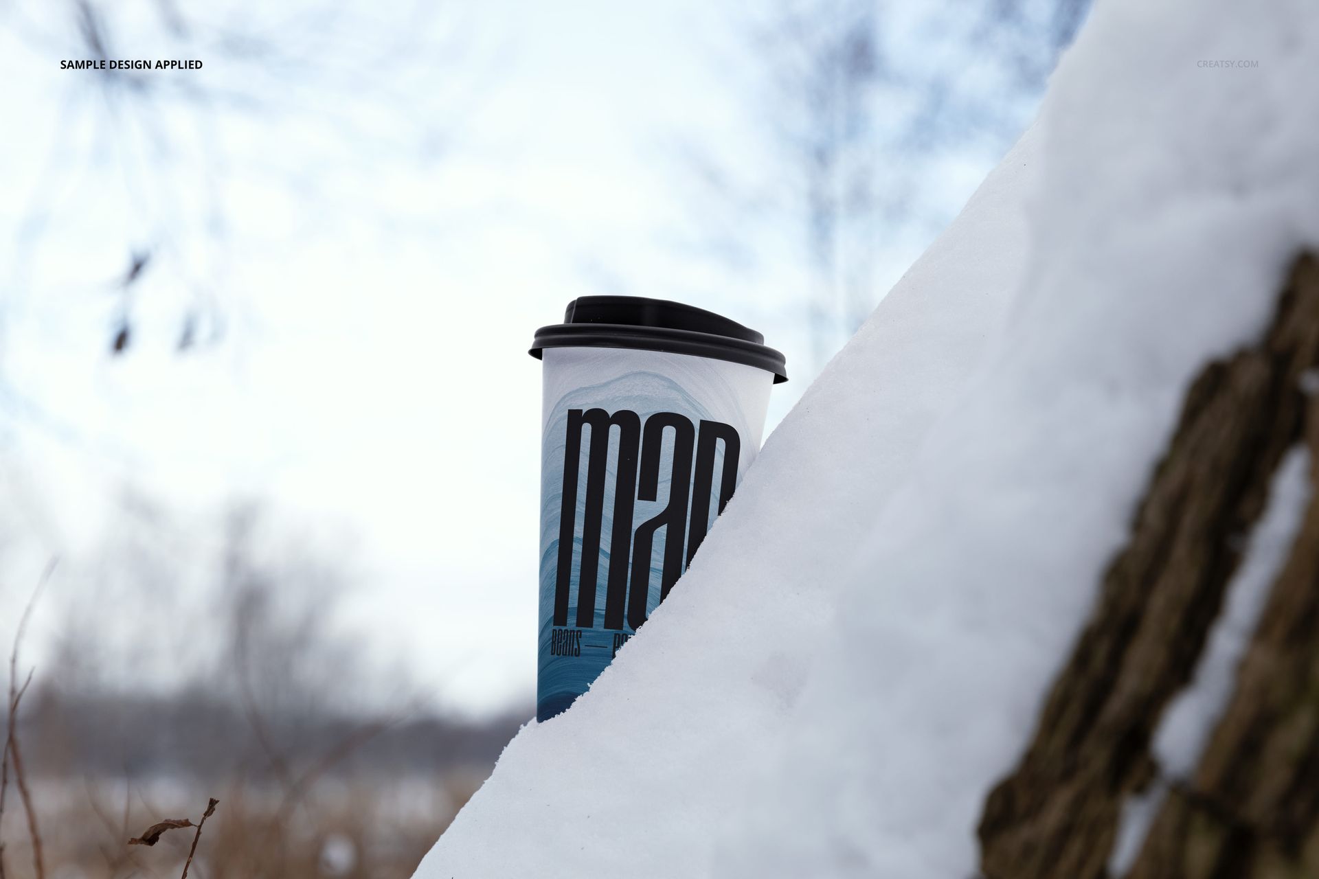 Winter scene with a paper cup mockup, black lid, and graphic pattern, placed beside a snowy tree trunk.