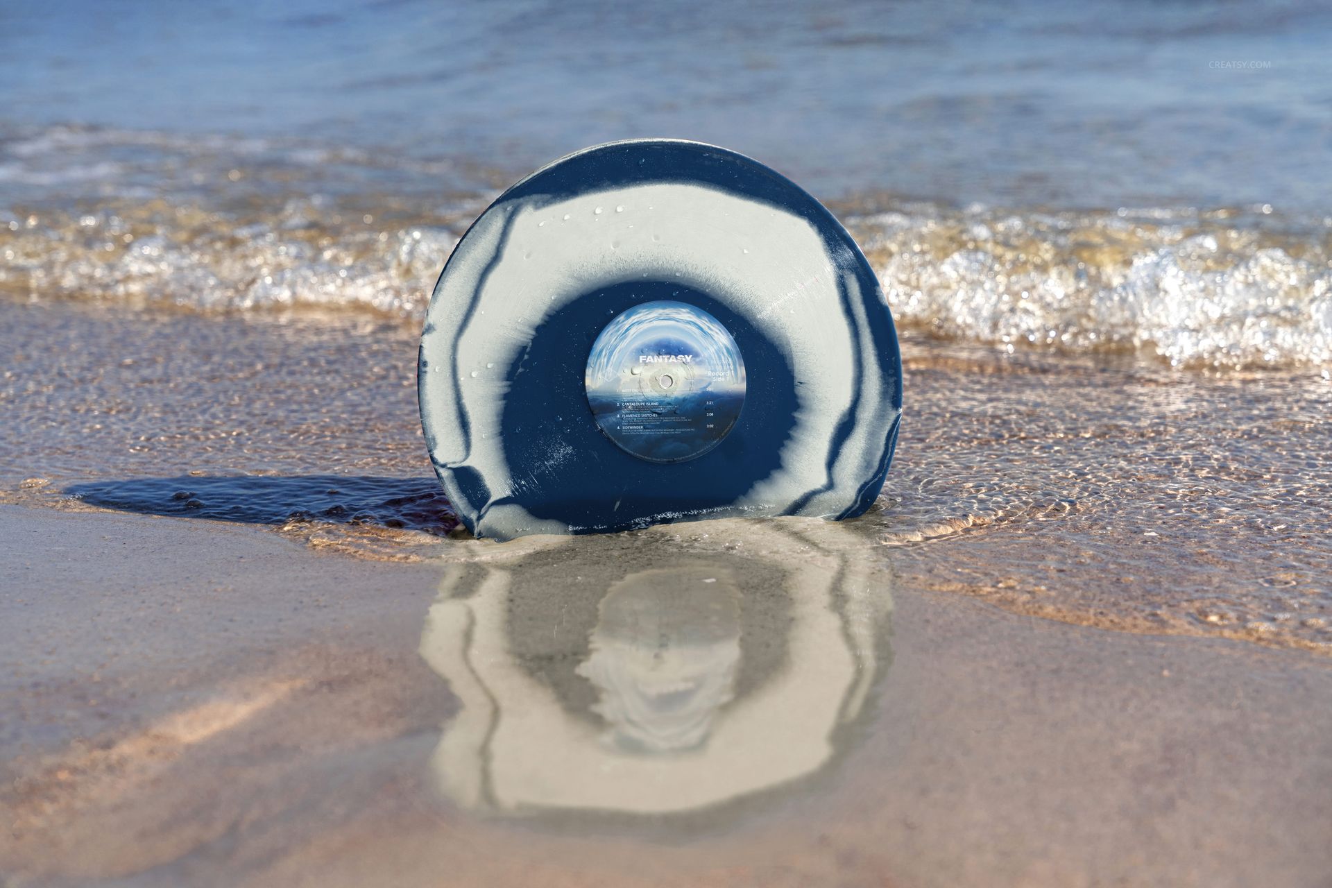 Blue and white vinyl record mockup resting on sandy beach with ocean waves in the background and reflection in water.
