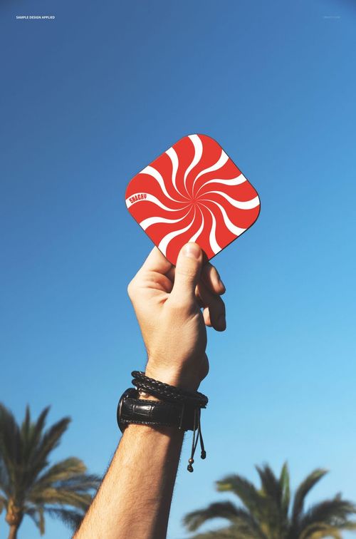 Person’s hand raised, displaying a mockup of a red and white patterned coaster with palm trees and sky in the background.