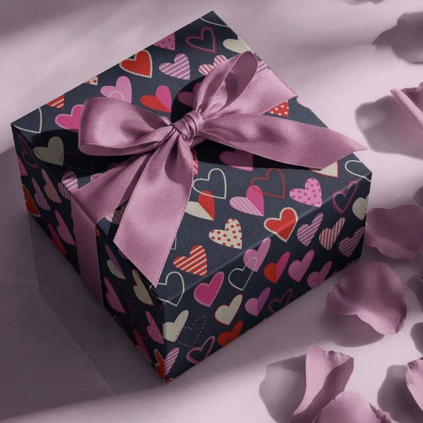Square gift box mockup with a purple ribbon, decorated with colorful heart patterns, surrounded by scattered pink petals.