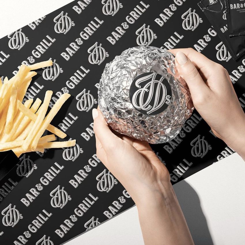 Hands holding a foil-wrapped burger with a round sticker on top, next to fries and branded packaging on a black background.