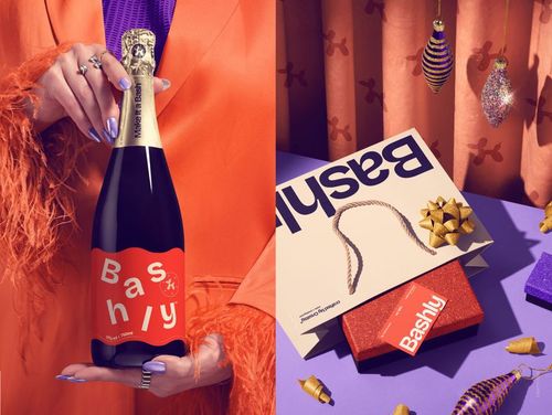 Front view of a person holding a champagne bottle with a red Bashly label, next to party-themed gift bags and boxes.