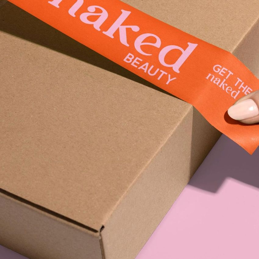 Packing tape mockup featuring orange tape with white text being applied to a plain brown cardboard box on a pink surface.
