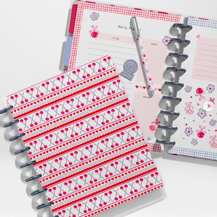 Top view of two spiral planners with colorful cherry and heart patterns, one open to a daily planner page with a pen on top.