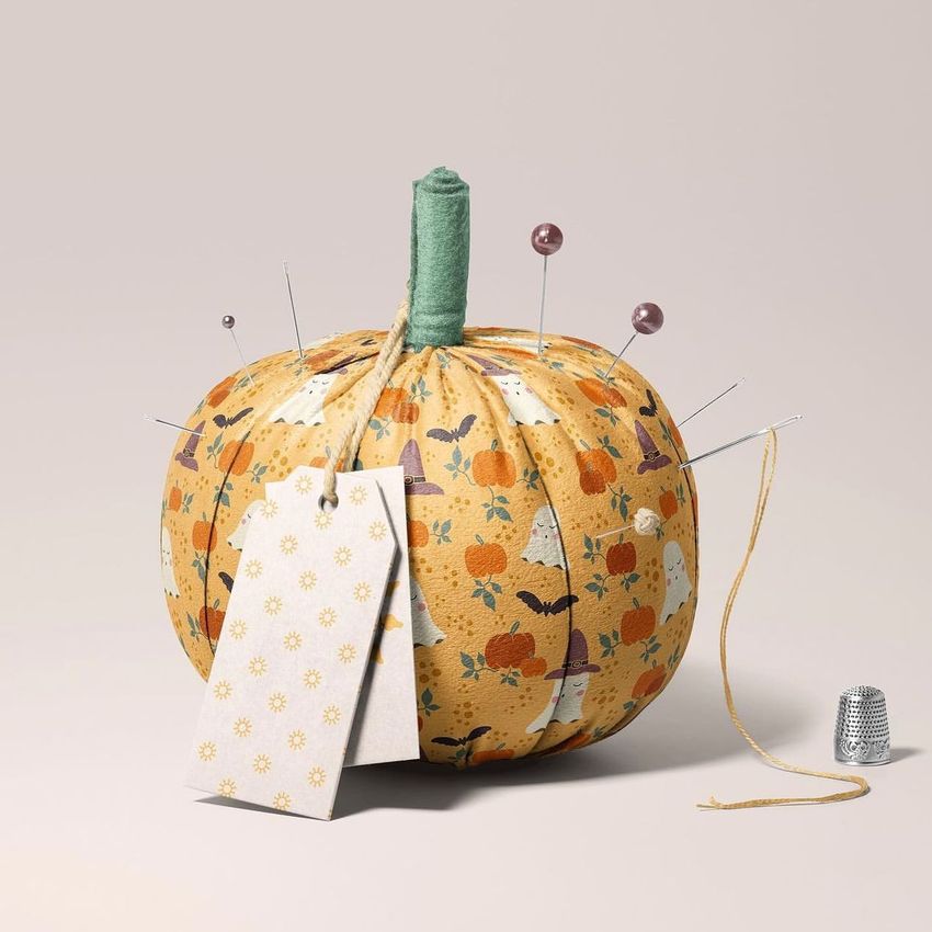 Fabric pumpkin mockup with a Halloween-themed pattern, sewing pins, a blank tag, and a thimble on a neutral background.