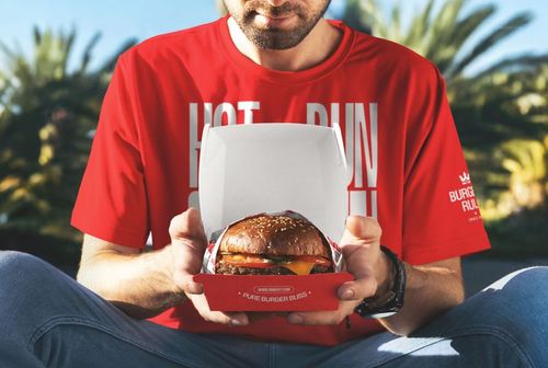 Front view of a cheeseburger in a mockup burger box held by a man in a red shirt, palm trees in background.