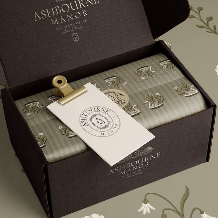 Mailing box mockup featuring an open dark box with patterned tissue paper and a branded tag clipped inside, set on a floral surface.