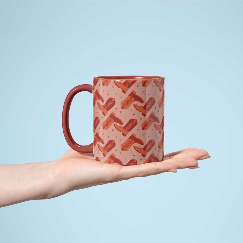 Sublimation mug mockup featuring a patterned coffee mug with a red handle, held on an open palm against a light blue background.