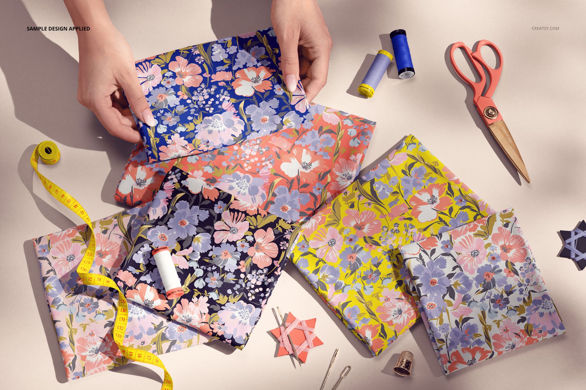 PSD mockup showing hands arranging floral patterned jersey fabrics, with scissors, thread, and tape measure on the table.