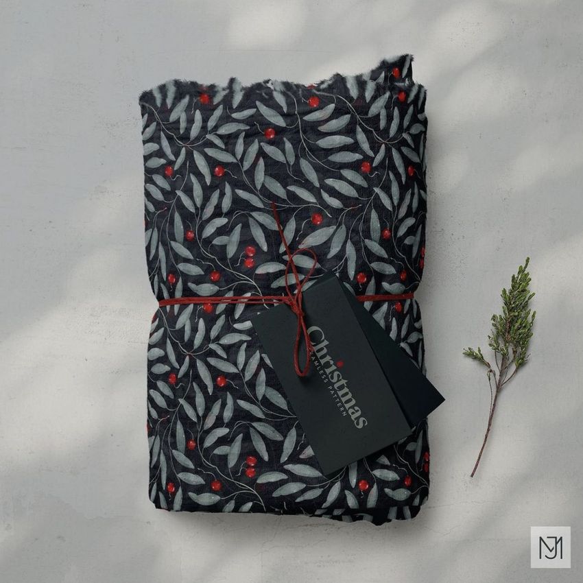 Muslin swaddle blanket mockup featuring a folded dark fabric with a festive leaf and berry pattern, tied with red string and a black tag.