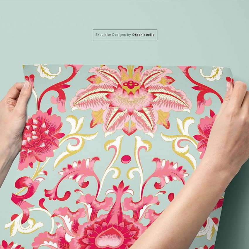 Hands holding a sheet of floral wallpaper mockup with large pink and red flowers on a light blue background, seen from above.