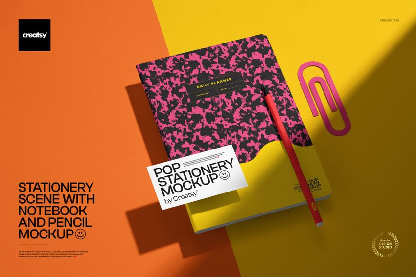Notebook and pencil mockup displayed from above on a split orange and yellow surface with a large pink paperclip prop.