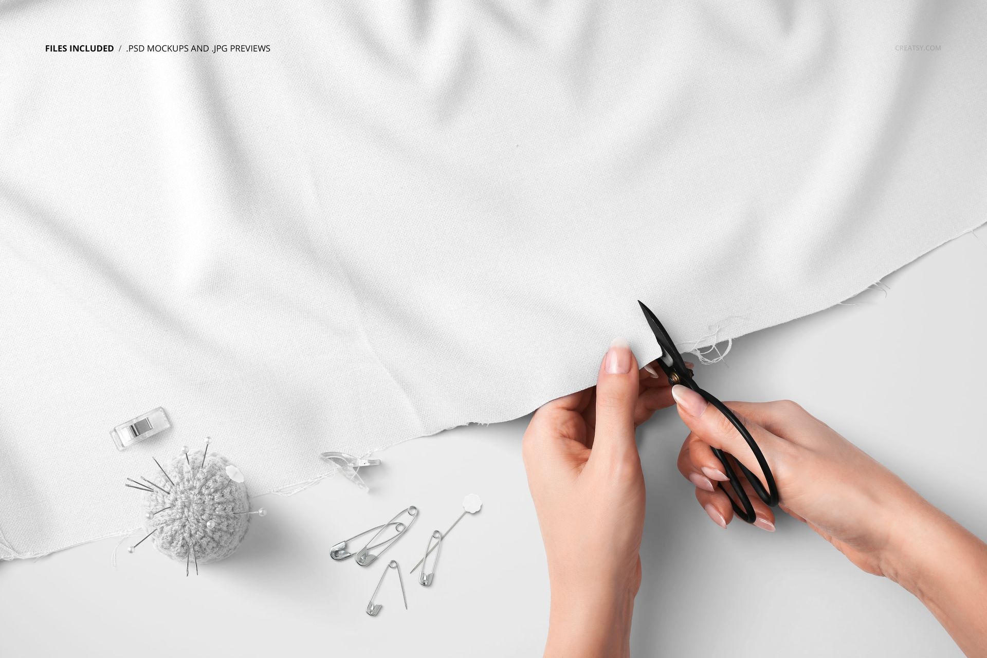 Top view of hands trimming white textile with scissors, next to a pincushion, safety pins, and a fabric mockup setup.