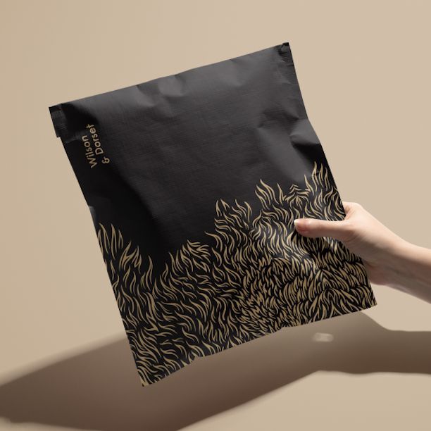 Hand holding a black cloth lined envelope mockup with gold abstract fur pattern and minimal text on a beige background.