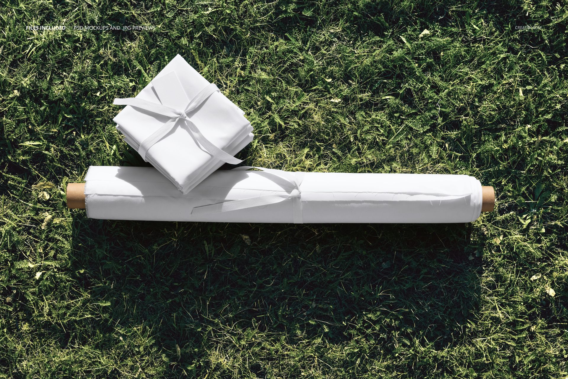 PSD mockup showing a fabric roll and folded fabric pieces with ties lying on a textured grass surface.