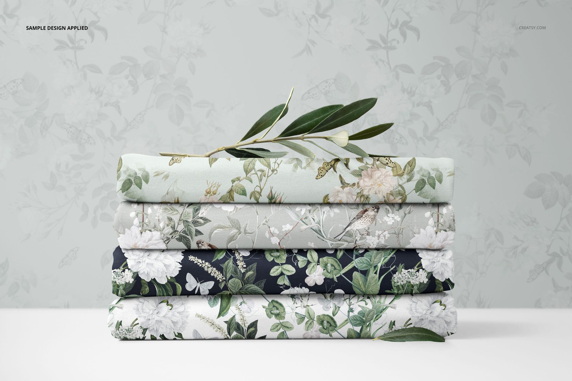 PSD mockup showing layered jersey fabrics in various botanical prints, stacked horizontally with olive branch accents on top.