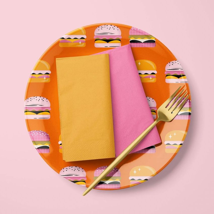 Napkin mockup set featuring a yellow and a pink napkin placed on an orange plate with a burger pattern, set on a pink background.