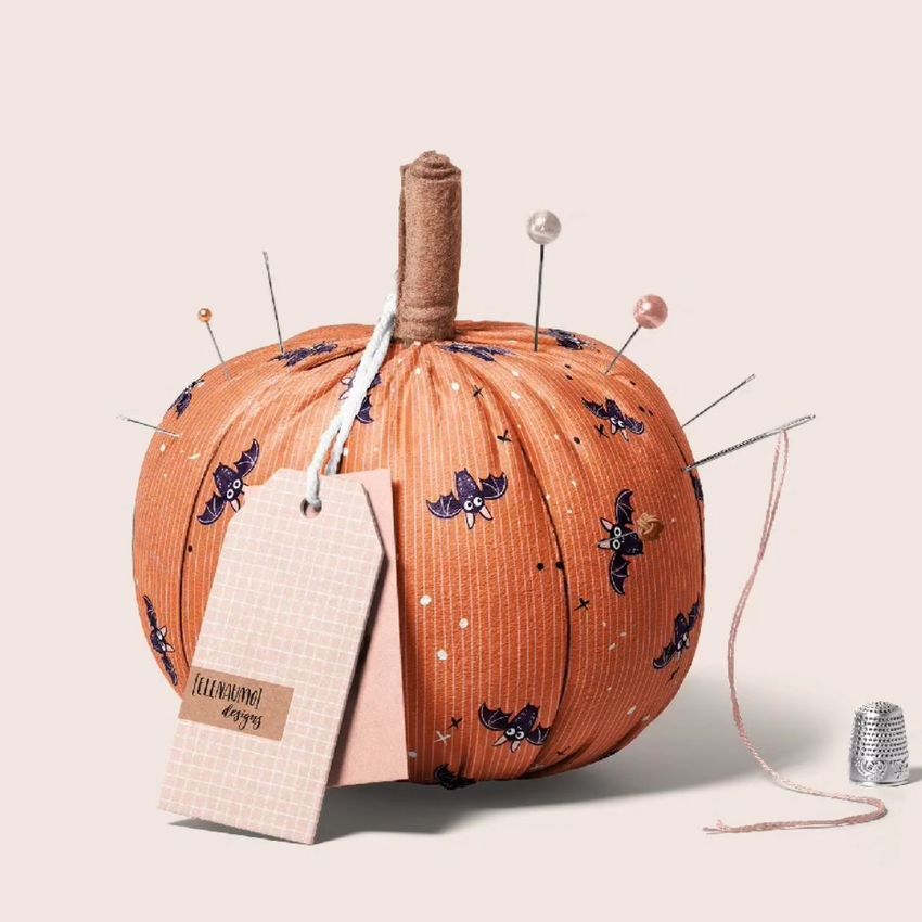 Fabric pumpkin mockup decorated with a Halloween bat pattern, pins, and tags, set on a light neutral background with a thimble.